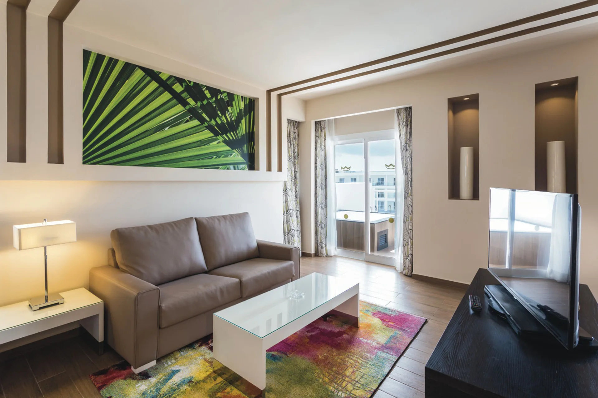 Suite living area with sofa, TV, balcony doors, and colorful rug at Hotel Riu Republica.