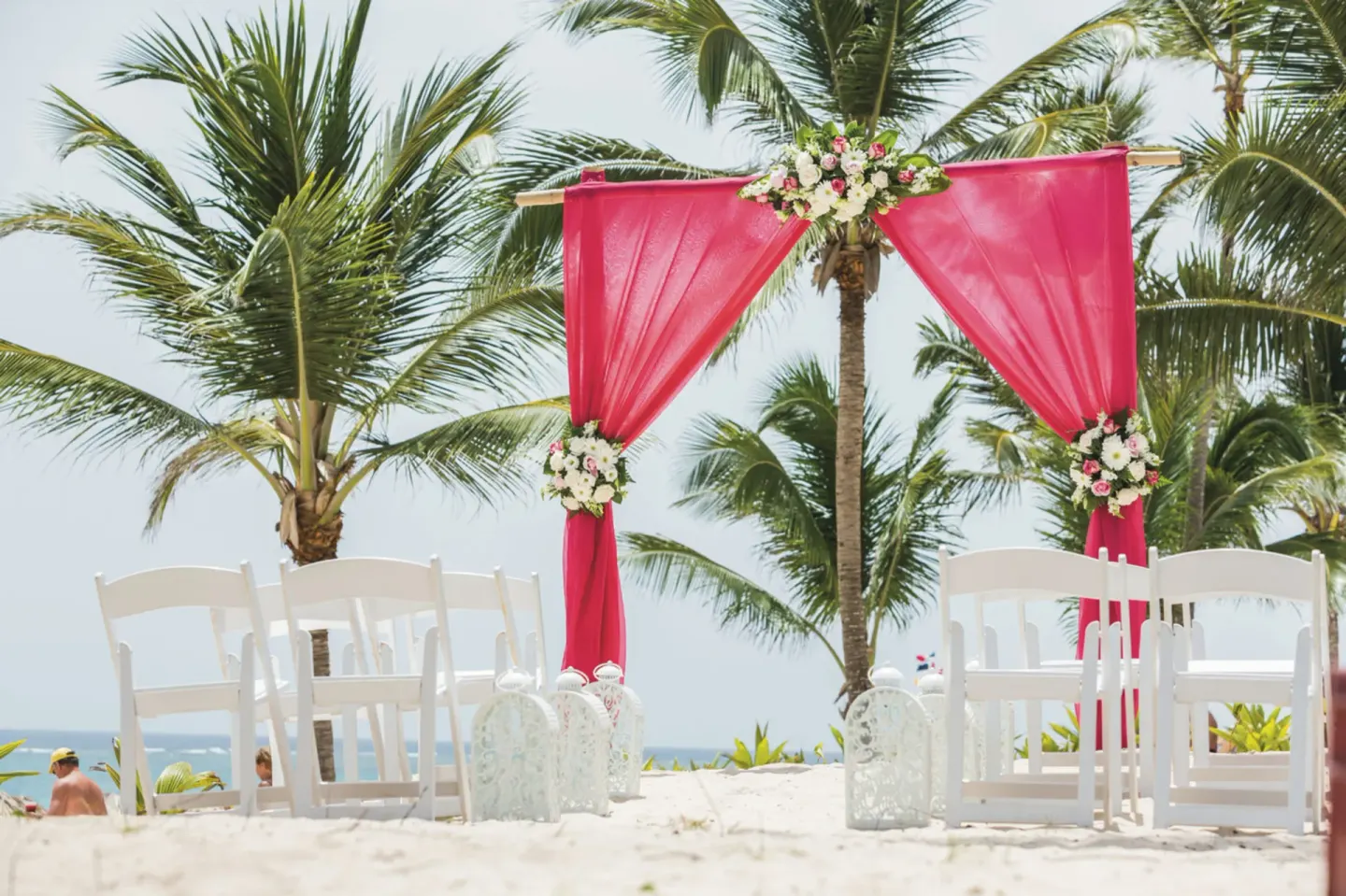 Pink beach wedding arch and white chairs among palms at Hotel Riu Republica.