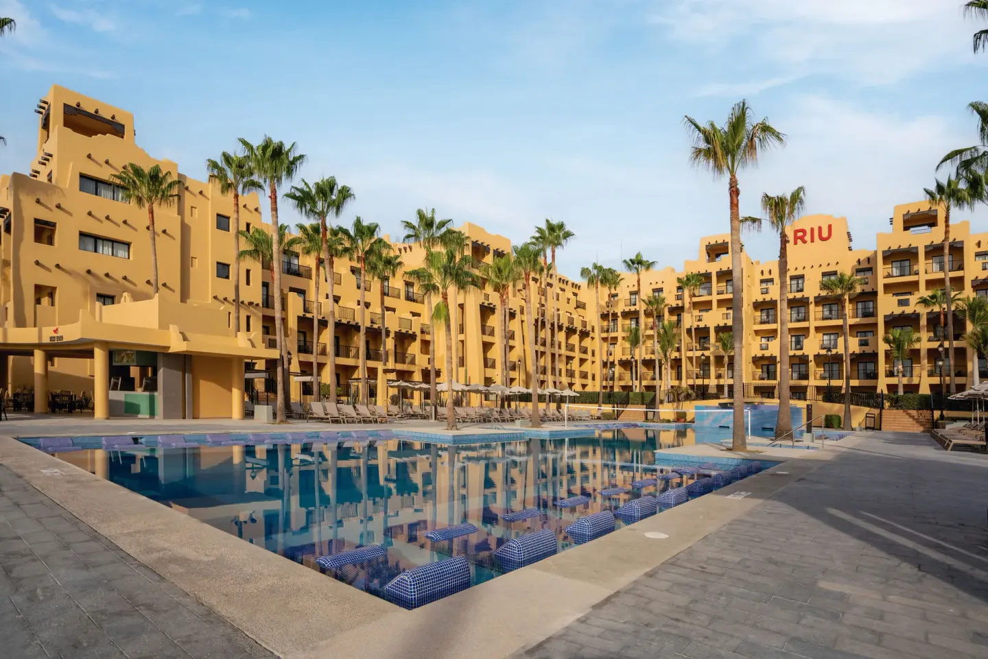 Large resort pool framed by palms and guestroom buildings at Hotel Riu Santa Fe.