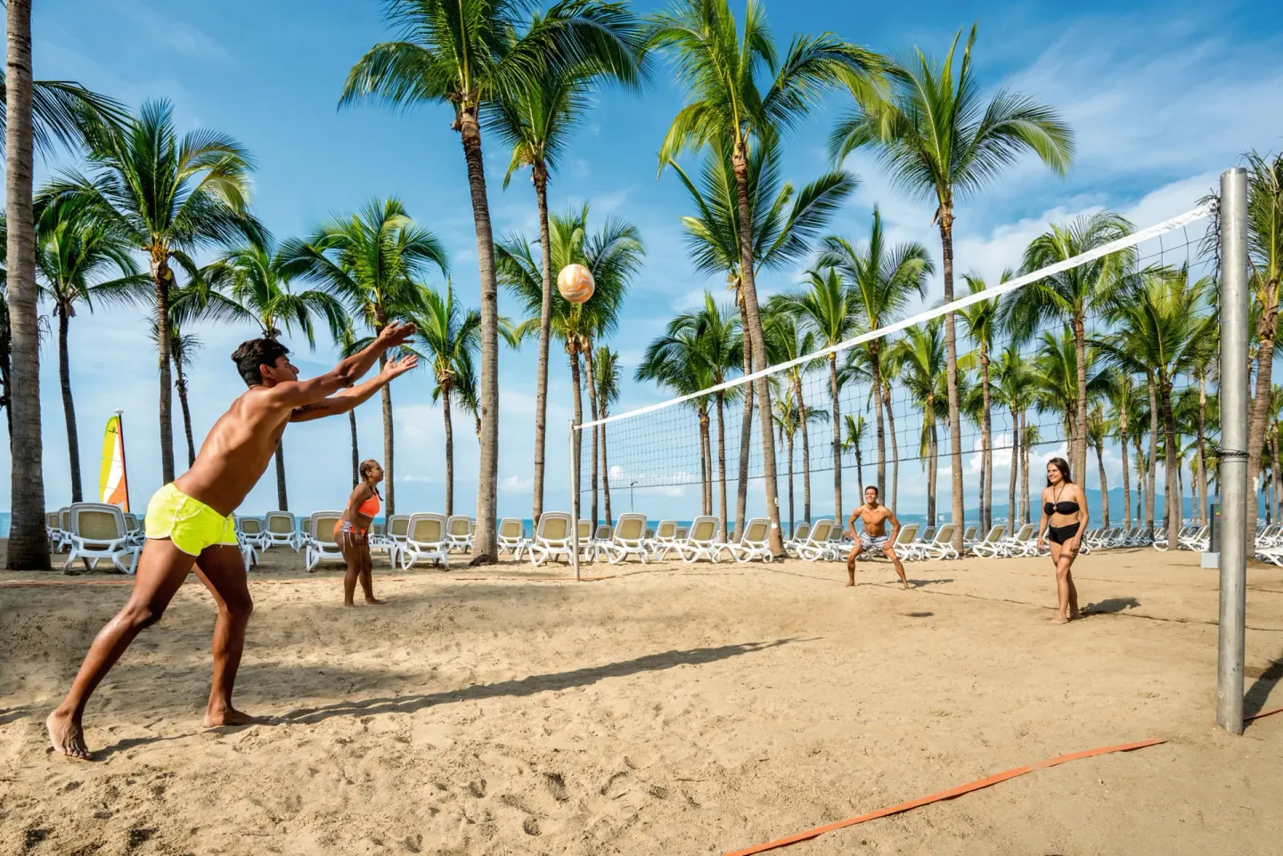 Guests play beach volleyball on sand beside palms and lounge chairs at Hotel Riu Vallarta.