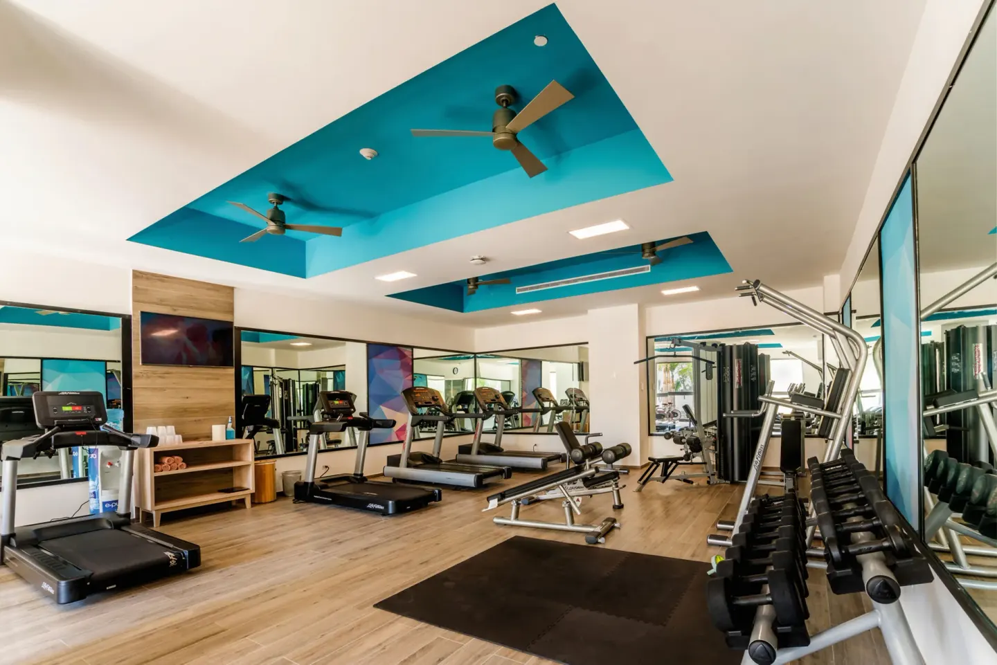 Fitness center with treadmills, weights, mirrors, and turquoise ceiling panels at Hotel Riu Vallarta.