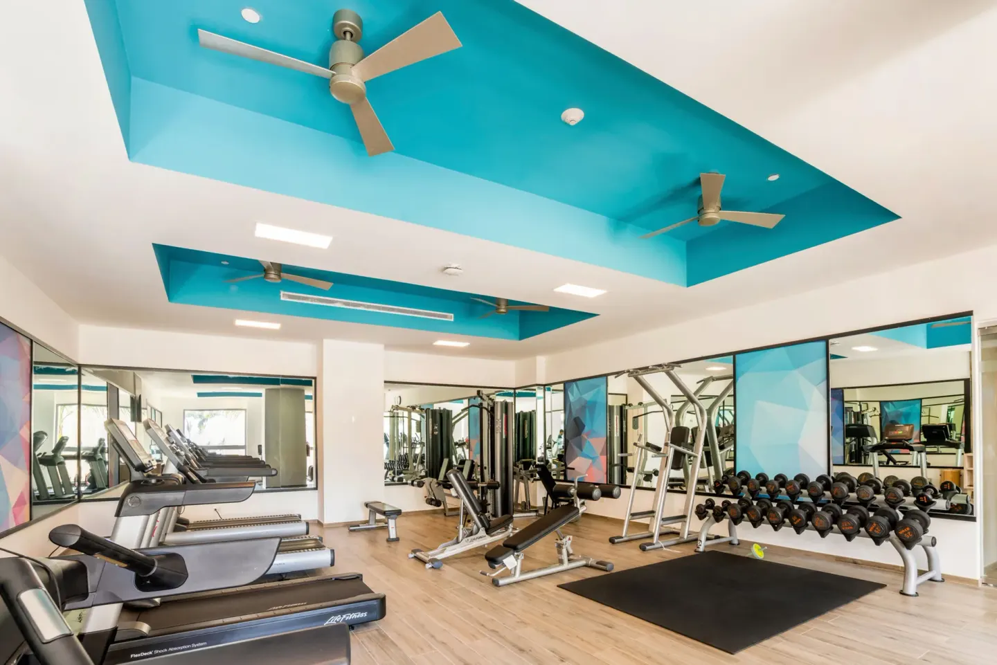 Hotel Riu Vallarta gym with treadmills, dumbbells, benches, mirrors, and turquoise ceiling accents.