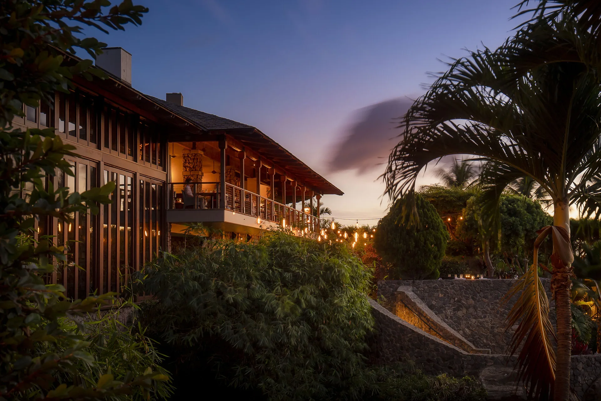A lit restaurant building sits among palms and gardens at dusk at Hotel Wailea.