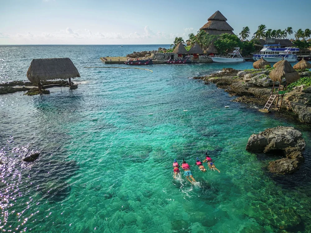 Snorkelers in clear coastal water near thatched buildings at Hotel Xcaret Mexico.