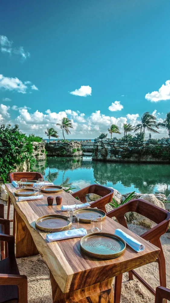 Wood dining table set beside a lagoon with rock walls and ocean beyond at Hotel Xcaret Mexico.