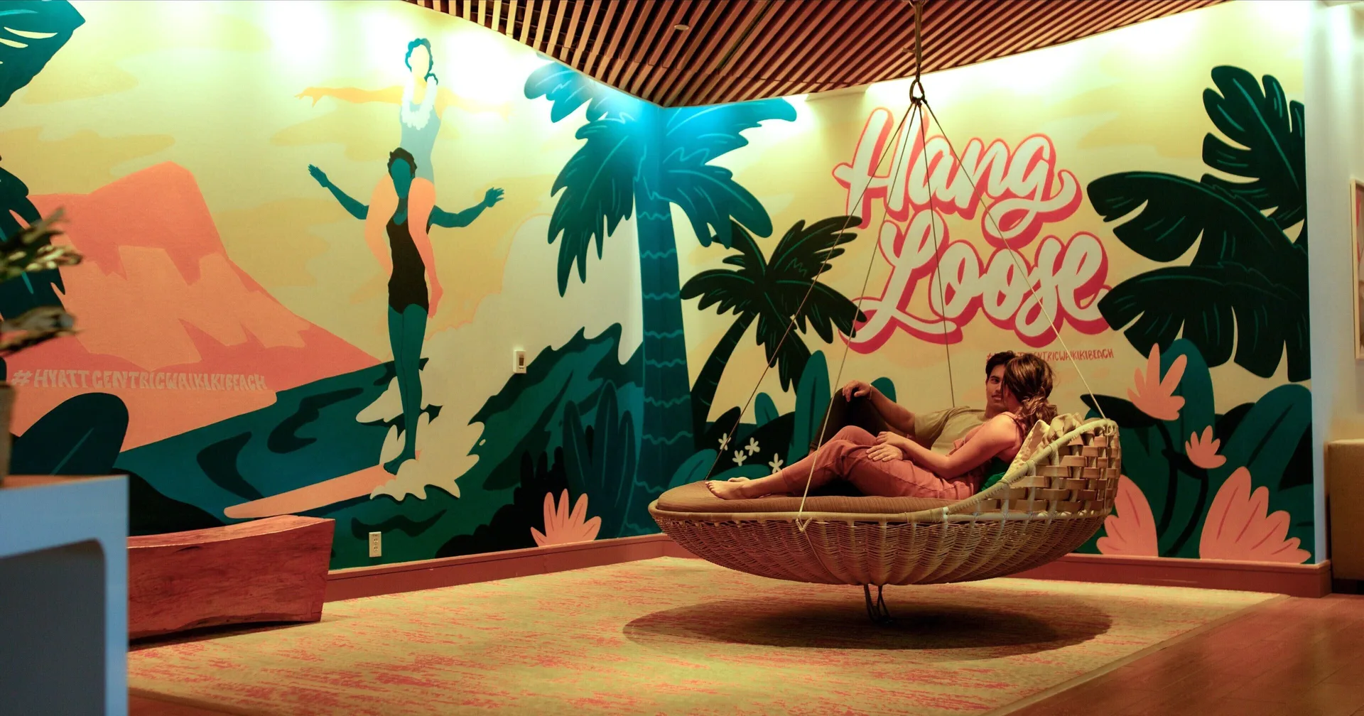 Guest relaxing in a hanging lounge chair beside a tropical mural at Hyatt Centric Waikiki Beach.