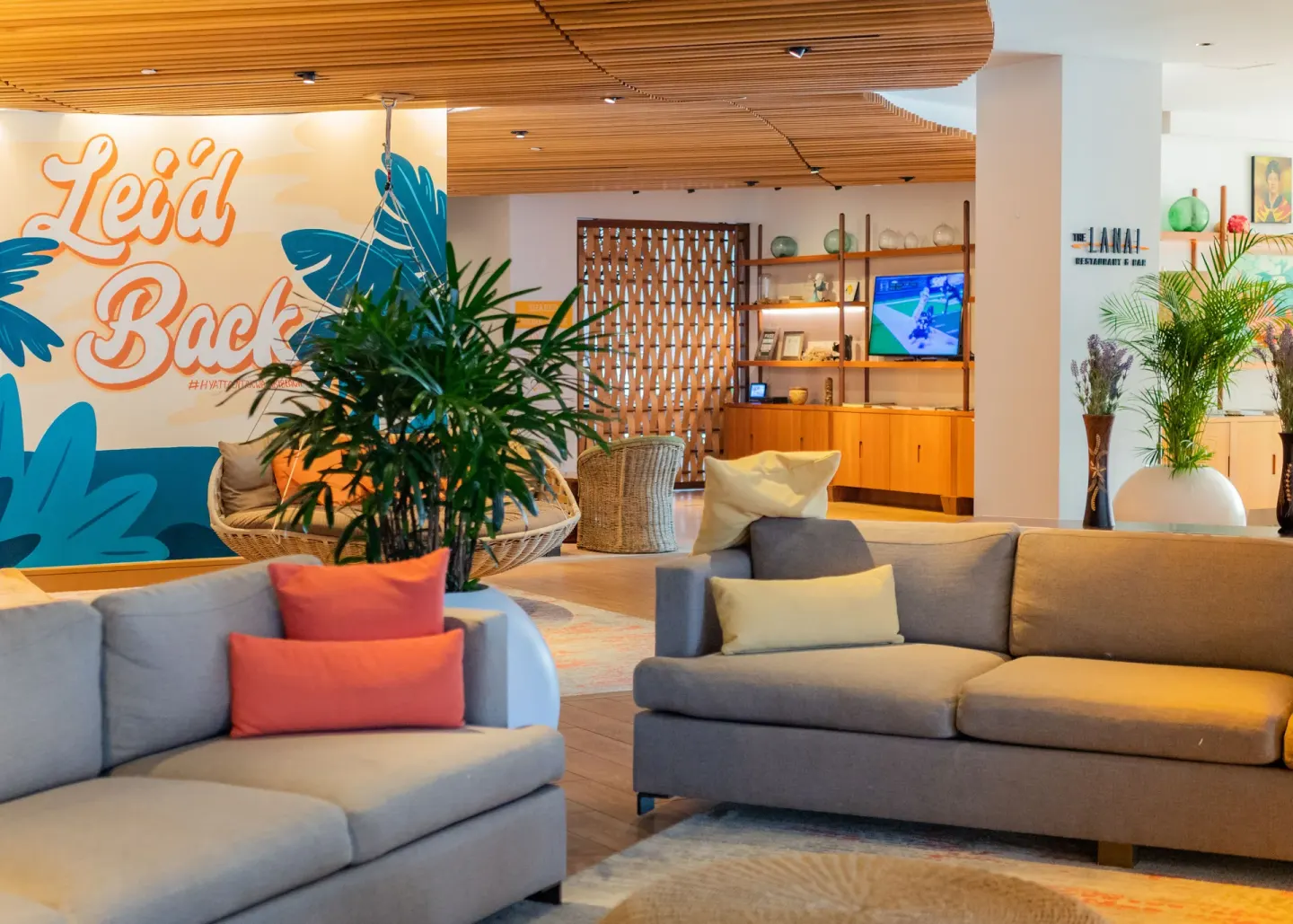Lobby lounge at Hyatt Centric Waikiki Beach with sofas, plants, mural, and wood ceiling details.