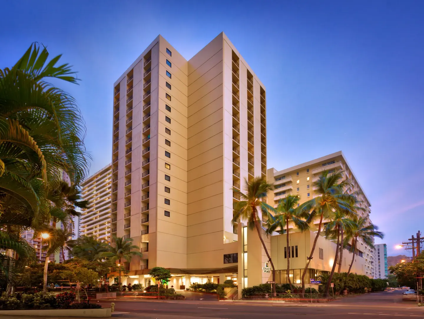 Tall hotel tower and palm-lined street at Hyatt Place Waikiki Beach.