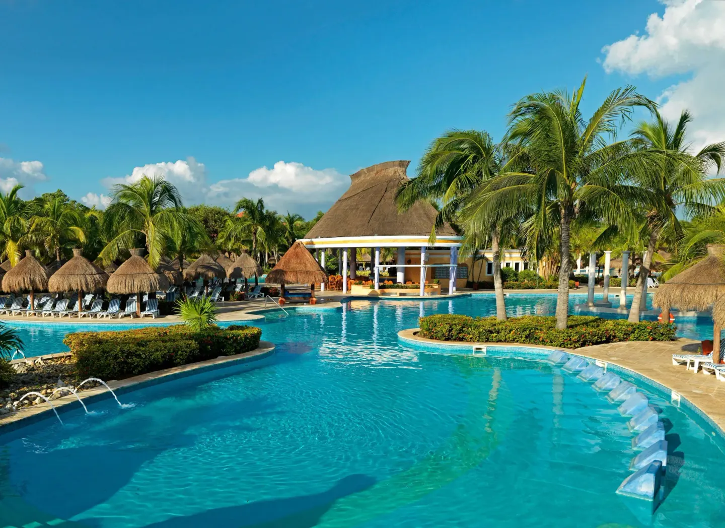 Resort pool with palapas, palms, and a thatched building at Iberostar Waves Paraíso Beach.