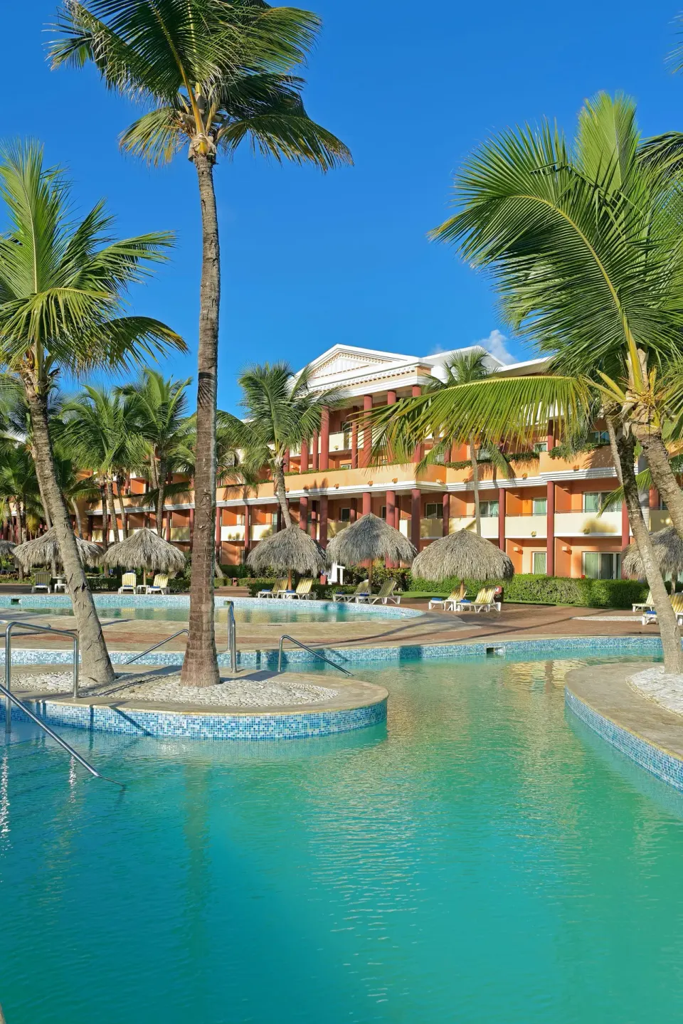 Pool with palm islands, palapas, and orange resort building at Iberostar Waves Punta Cana.