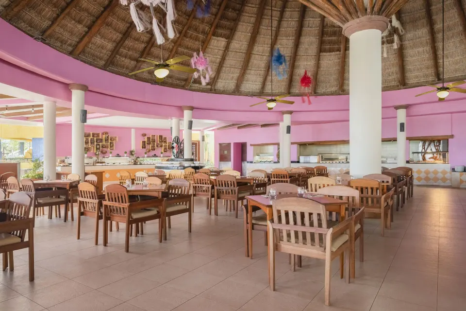 Pink-painted circular dining hall with wooden chairs under a thatched roof at Iberostar Waves Tucán