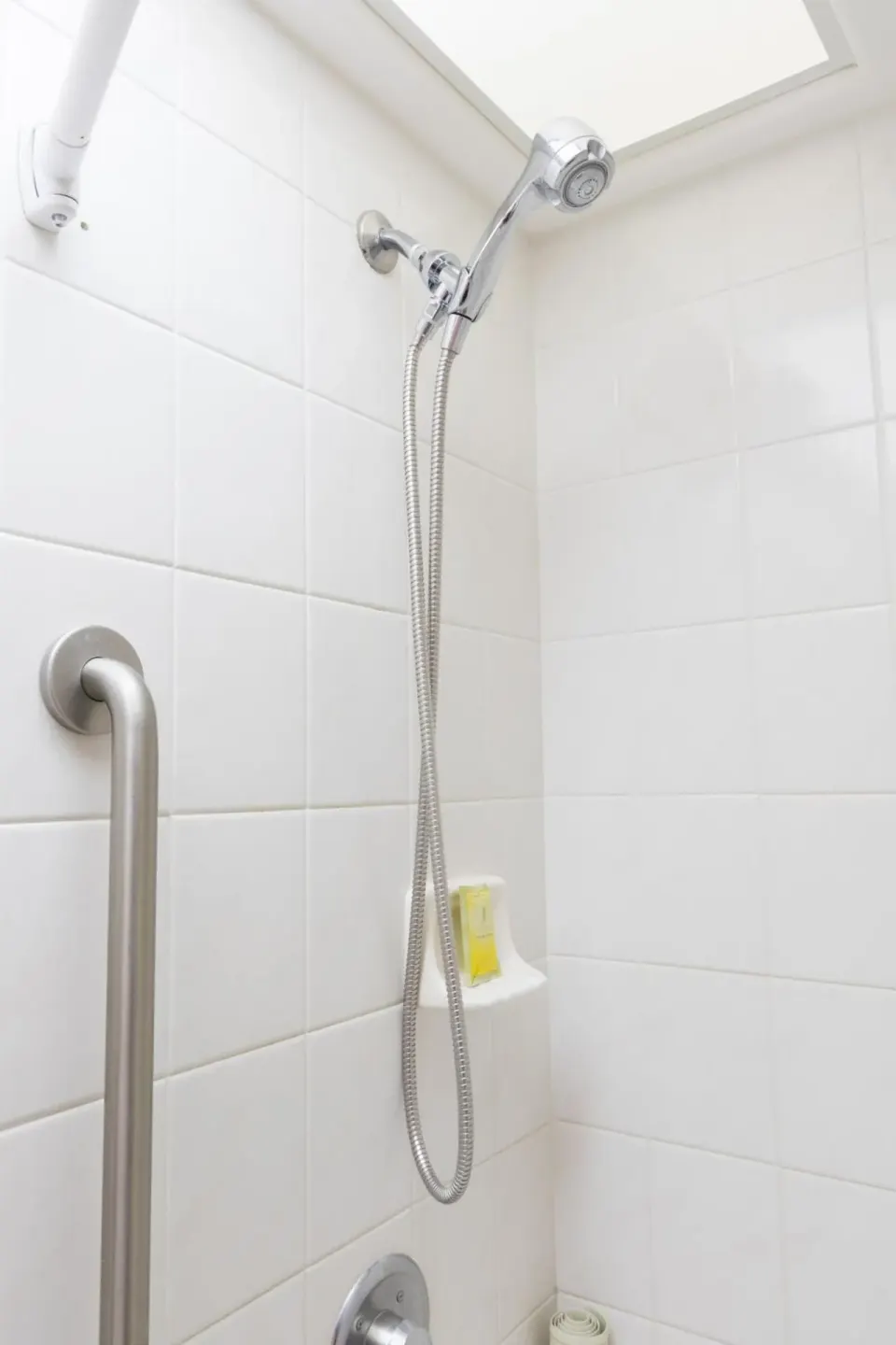 White tiled shower at Ilima Hotel with handheld showerhead and grab bar.