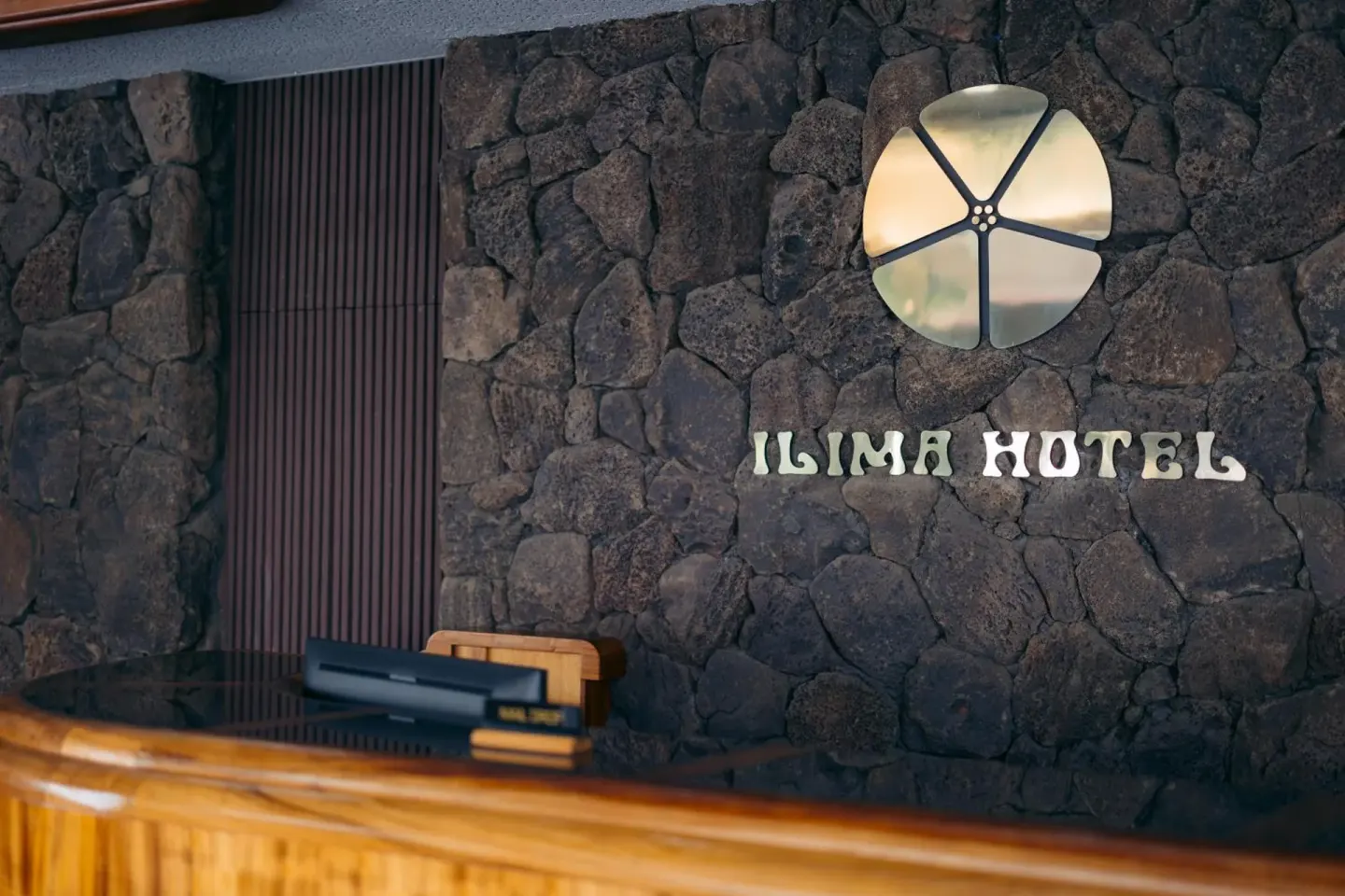 Lobby desk with stone wall and Ilima Hotel sign behind it.
