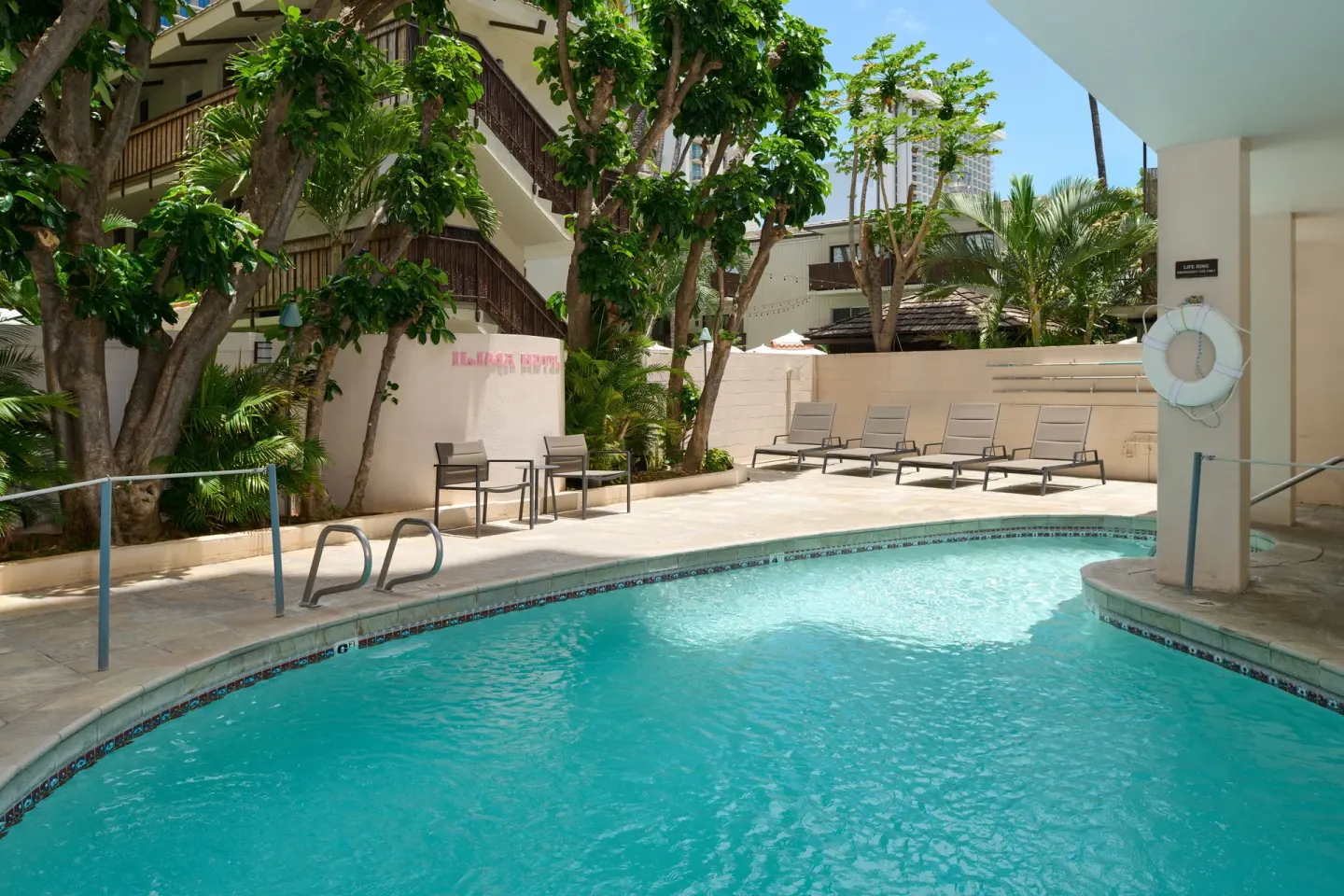 Outdoor pool with lounge chairs, trees, and railings at Ilima Hotel.