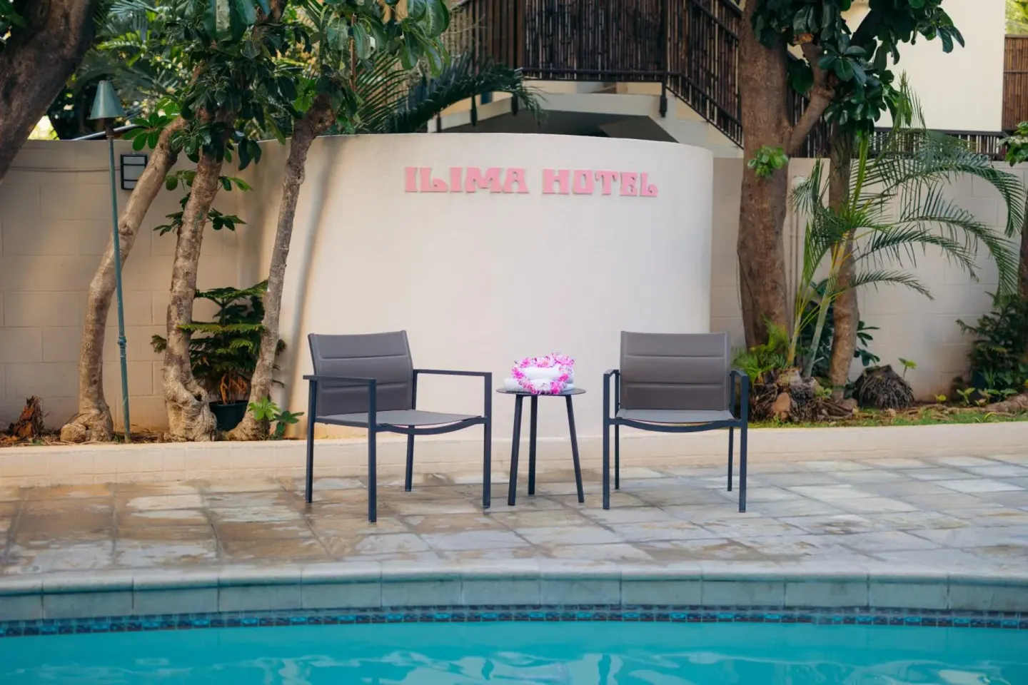 Poolside seating area with Ilima Hotel sign, chairs, table, and tropical plants.