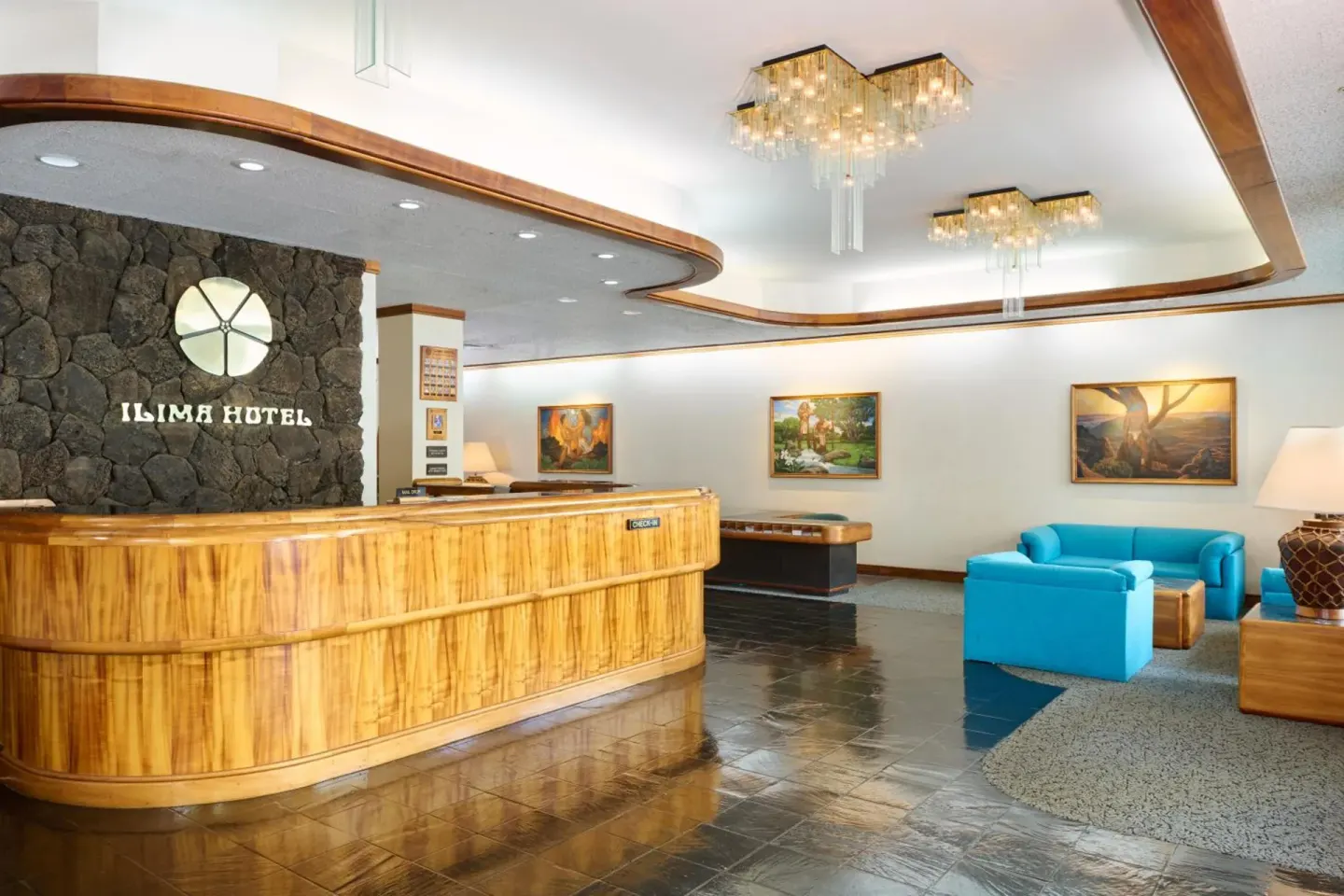 Wide lobby view at Ilima Hotel with reception desk, chandeliers, artwork, and blue chairs.