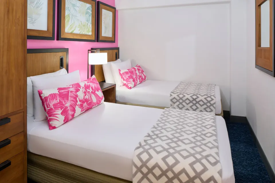 Suite bedroom with two beds, pink pillows, wall art, and a mirror at Ilima Hotel.