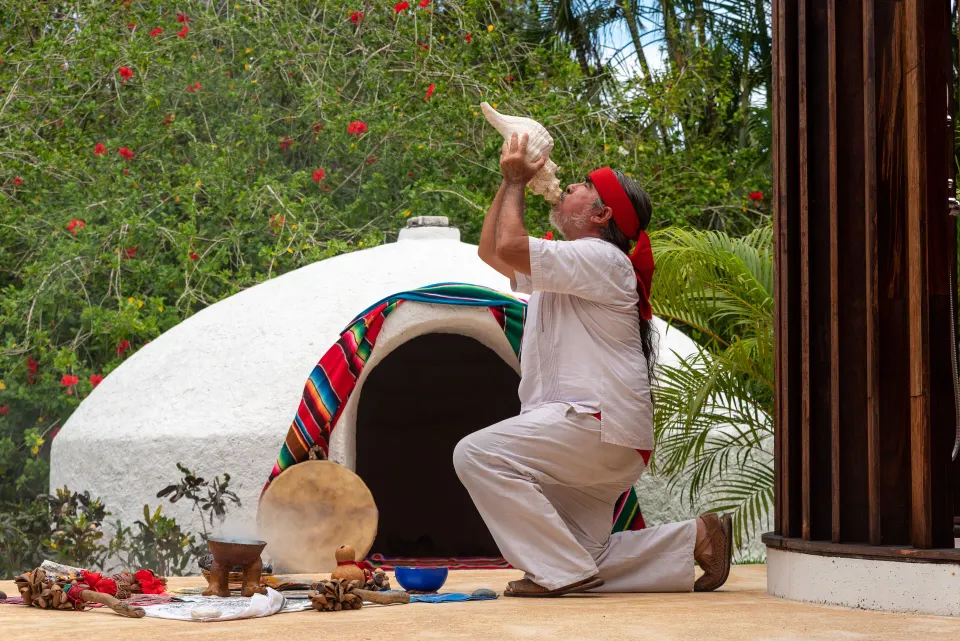 Performer kneeling and blowing a conch shell in outdoor ritual at InterContinental Presidente Cozumel Resort Spa