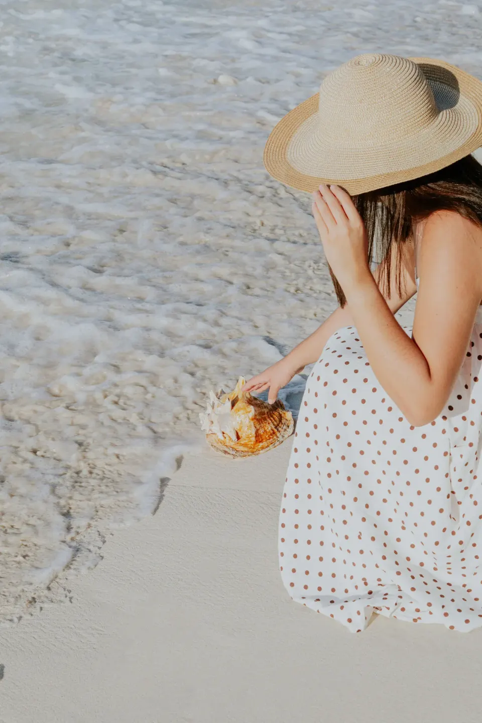 Woman in a sun hat holds a seashell at the shoreline by Ixchel Beach Hotel.