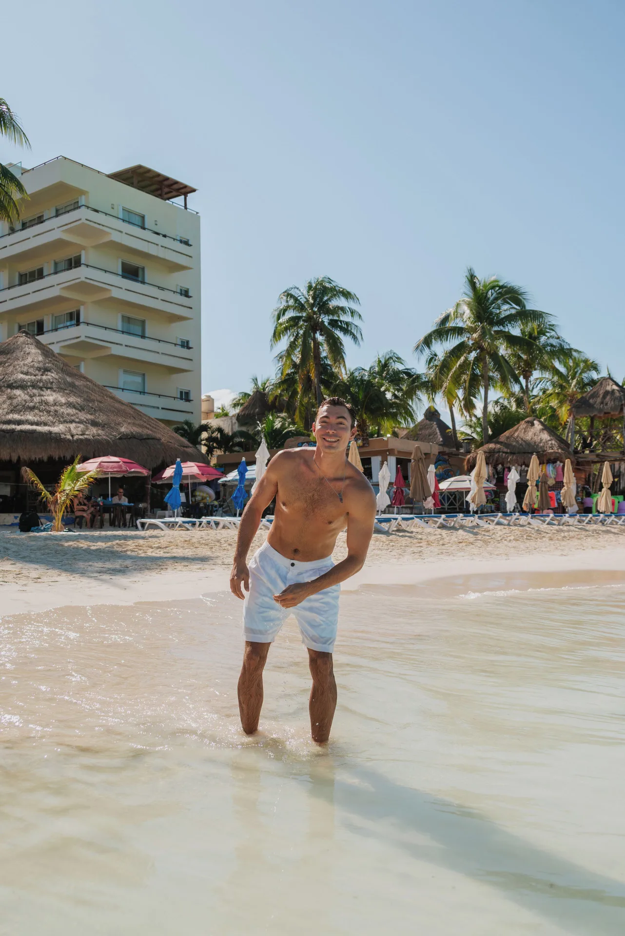 Man stands in shallow water with Ixchel Beach Hotel buildings and palms behind him.