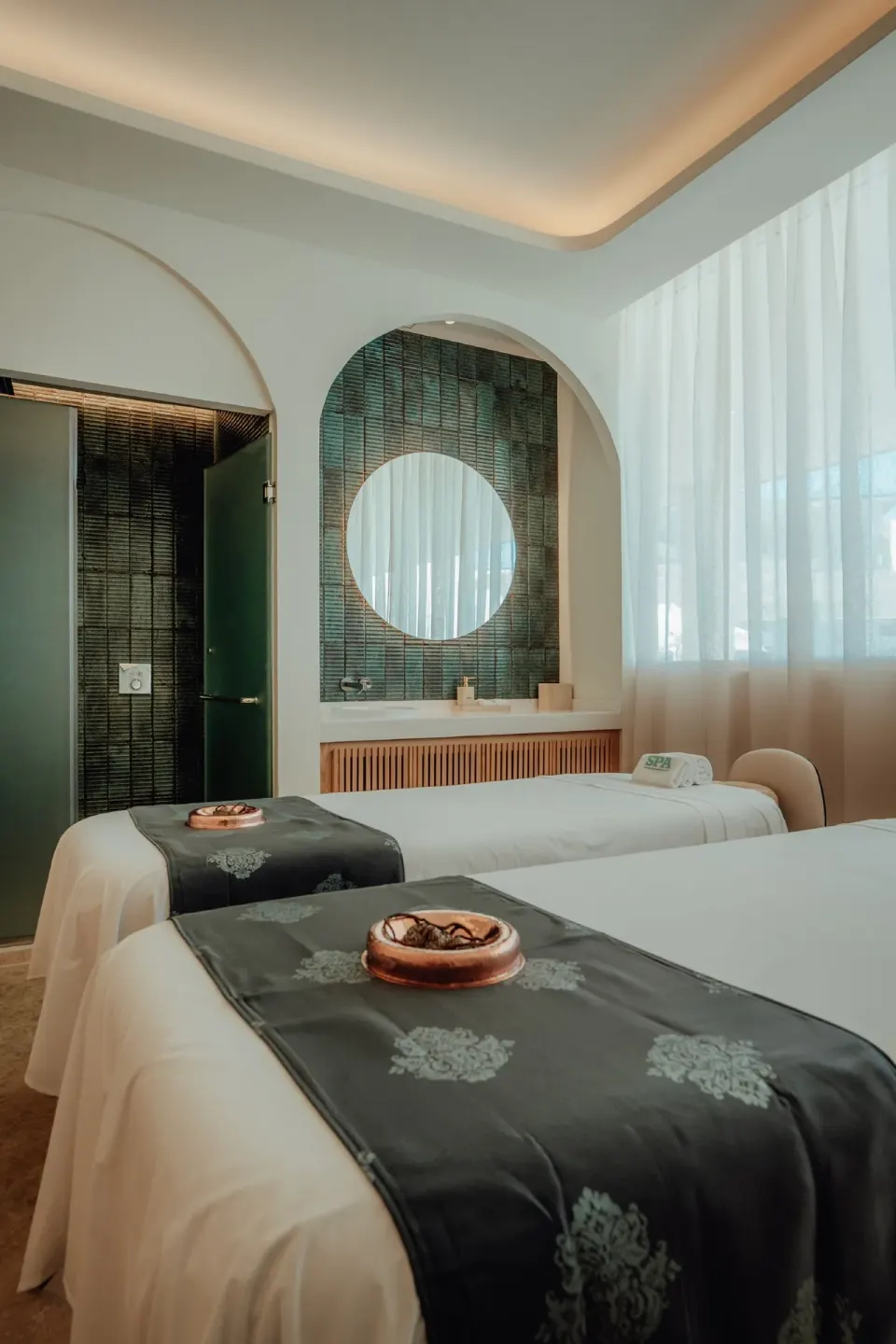 Two spa treatment beds with mirrors, shower area, and sheer curtains at JOIA Aruba.