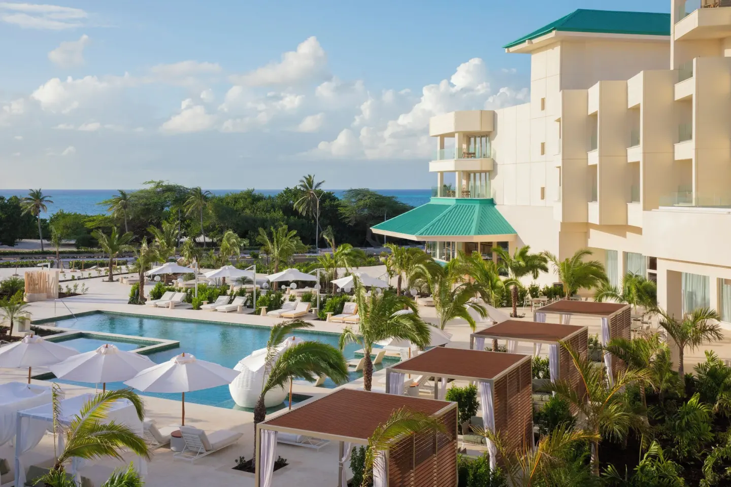 Pool, cabanas, palms, resort building, and ocean beyond at JOIA Aruba.