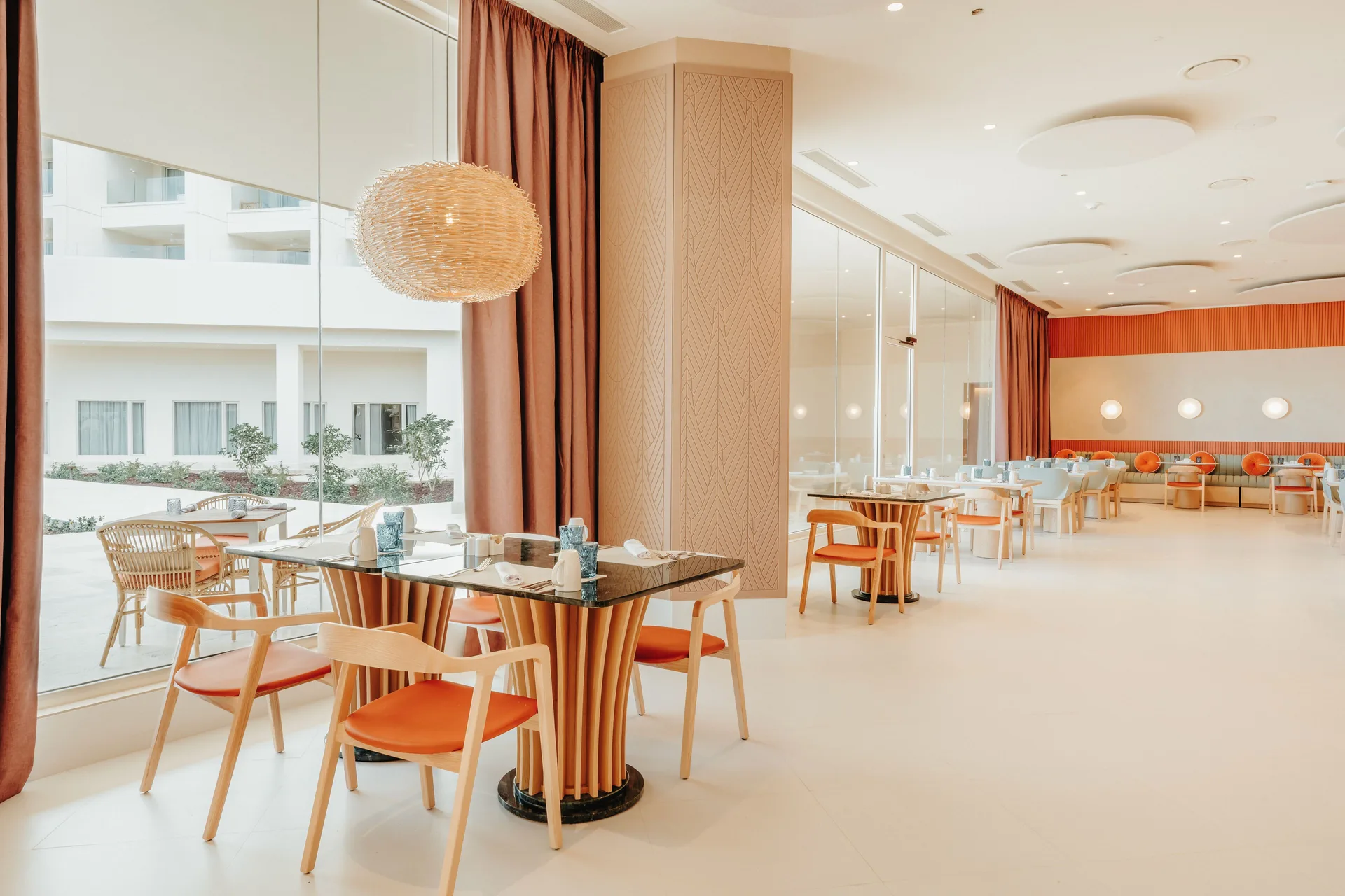 Bright restaurant seating with orange chairs and large windows at JOIA Aruba.