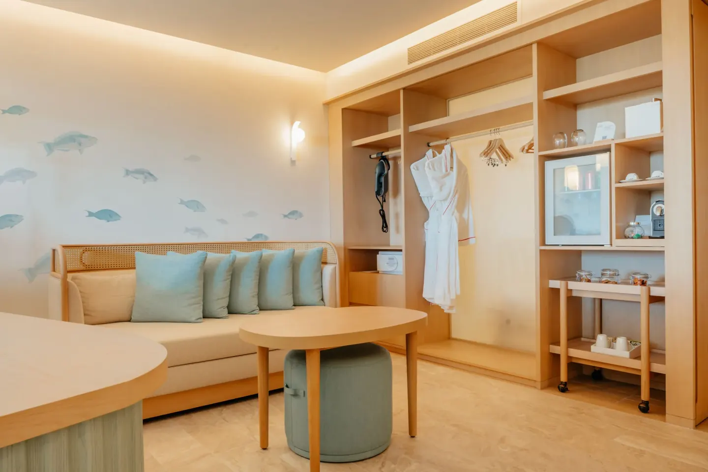 Suite sitting area with sofa, open wardrobe, robes, and refreshment cart at JOIA Aruba.