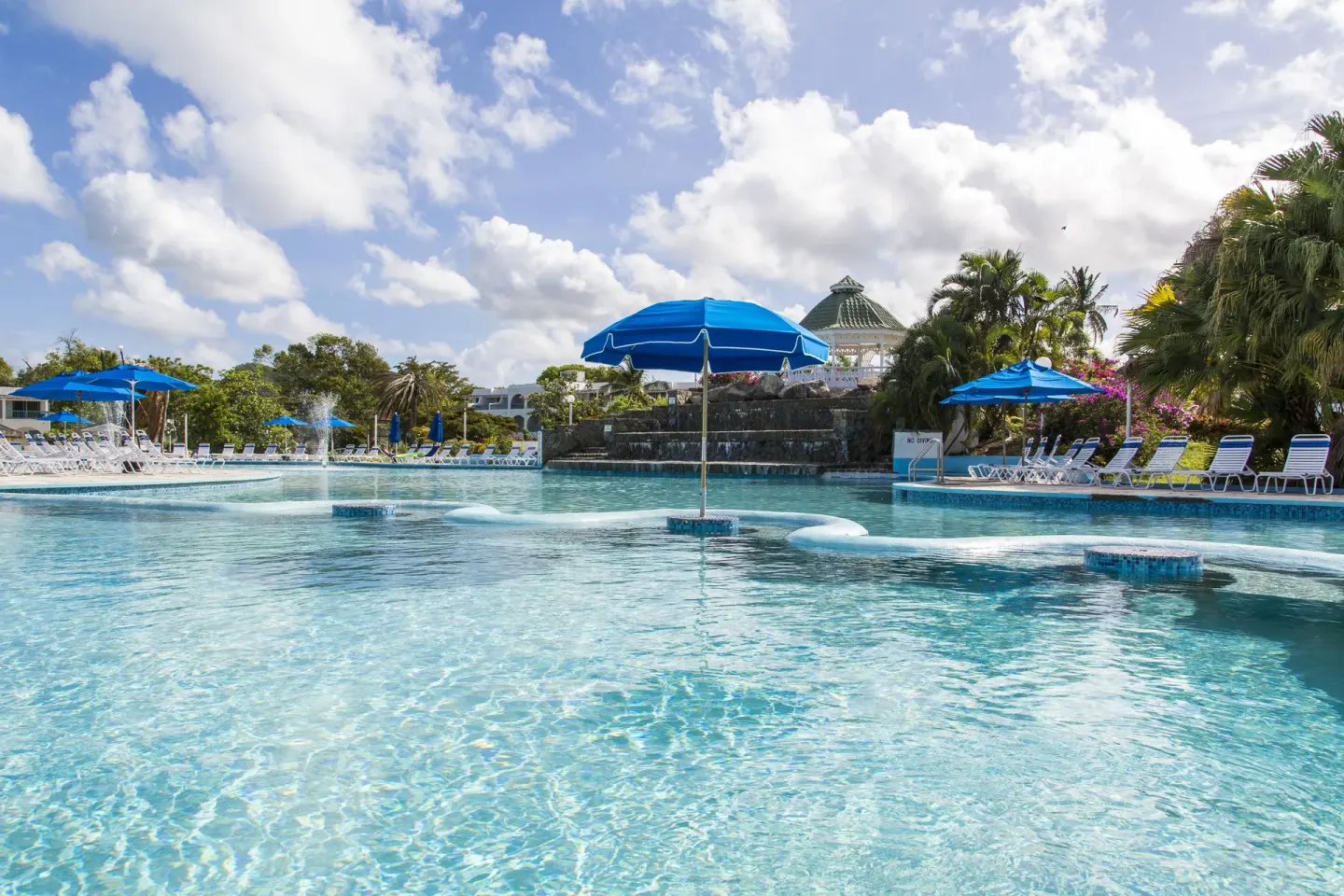 Resort pool with blue umbrellas, loungers and rock waterfall feature at Jolly Beach Antigua.