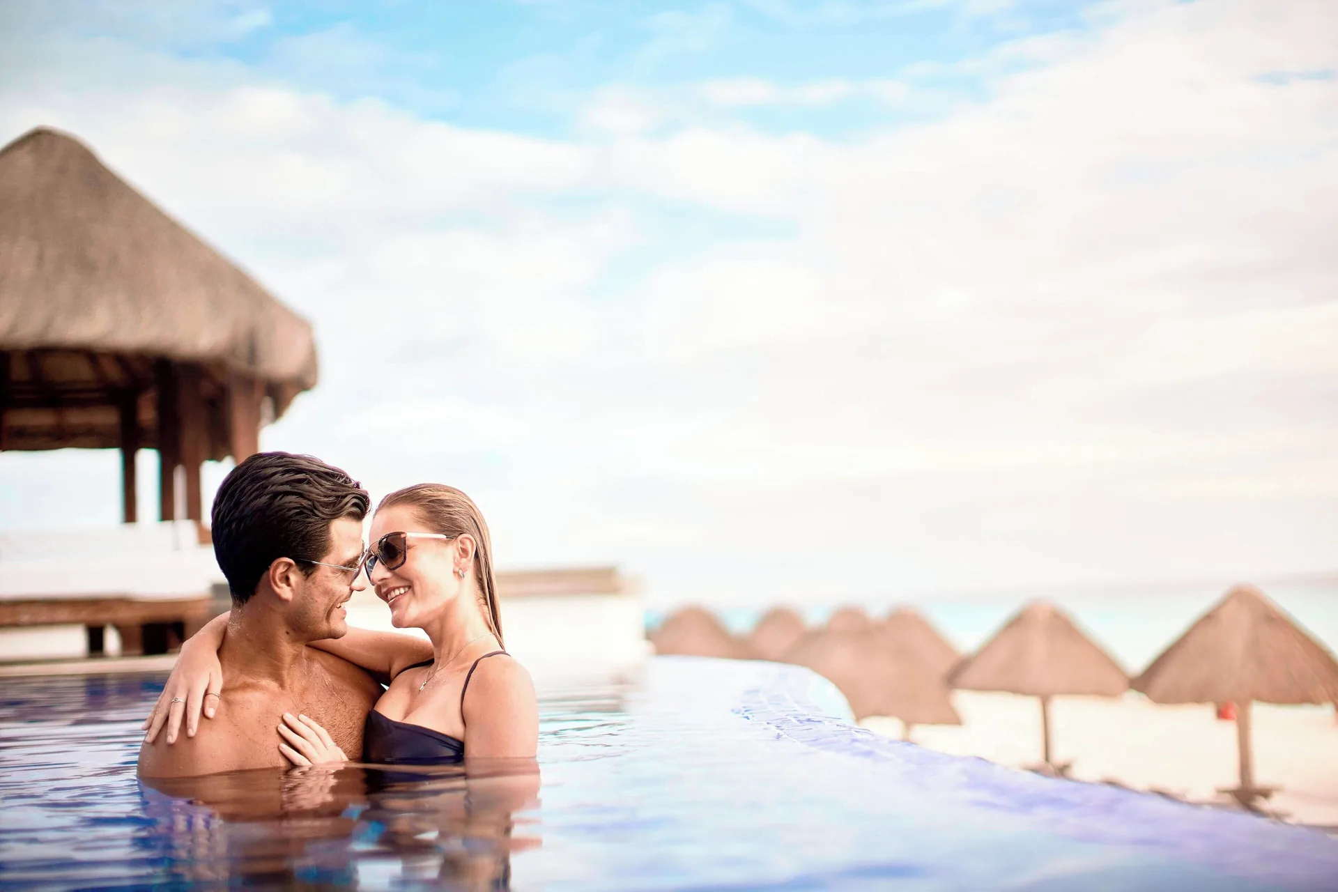 Couple embracing in an infinity pool at JW Marriott Cancun Resort & Spa with beach palapas beyond.