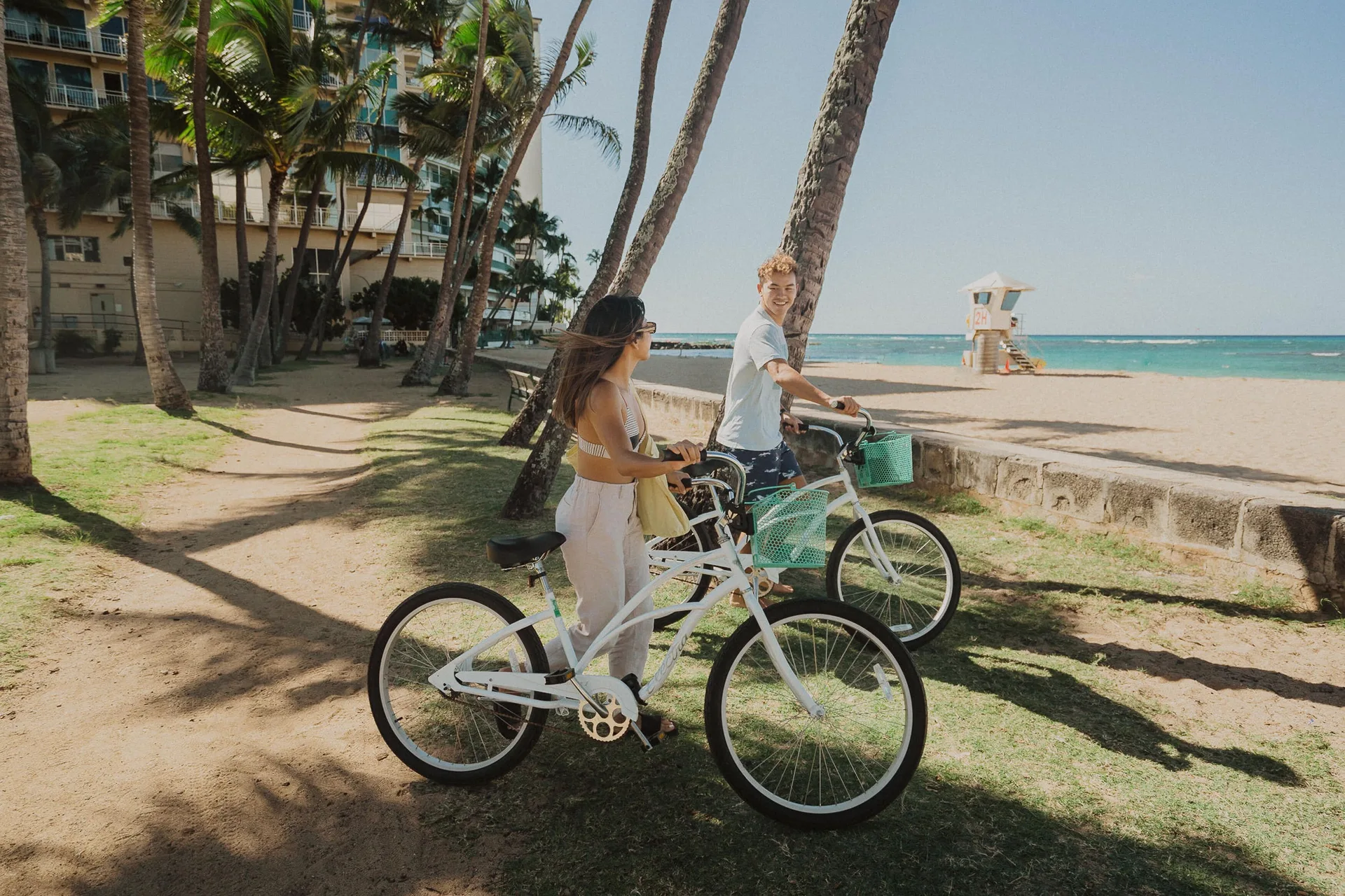 Two people with bicycles under palm trees by the beach at Kaimana Beach Hotel.