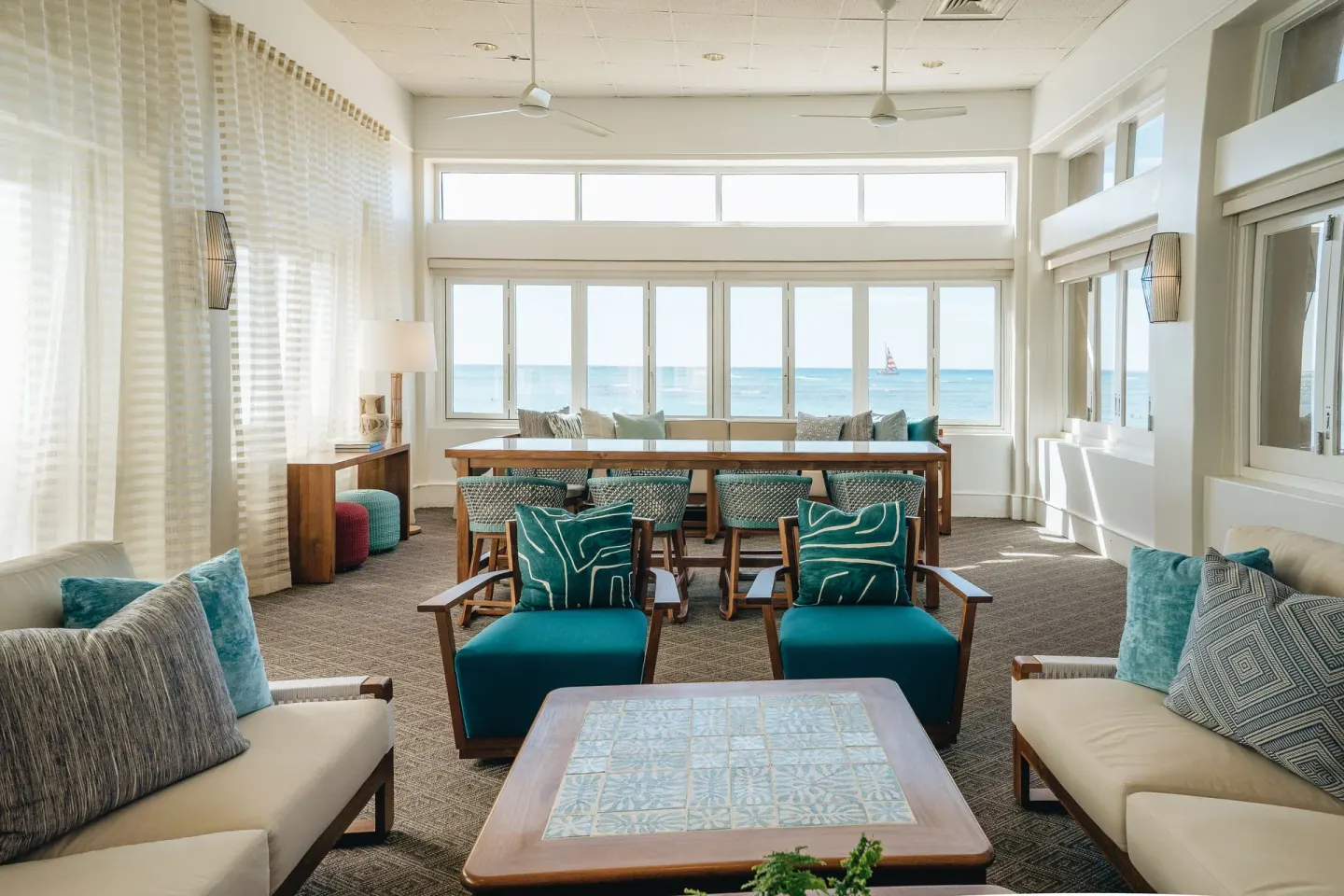 A bright lounge with ocean-facing windows and teal seating is shown at Kaimana Beach Hotel.