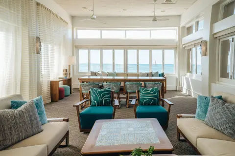 A bright lounge with ocean-facing windows and teal seating is shown at Kaimana Beach Hotel.