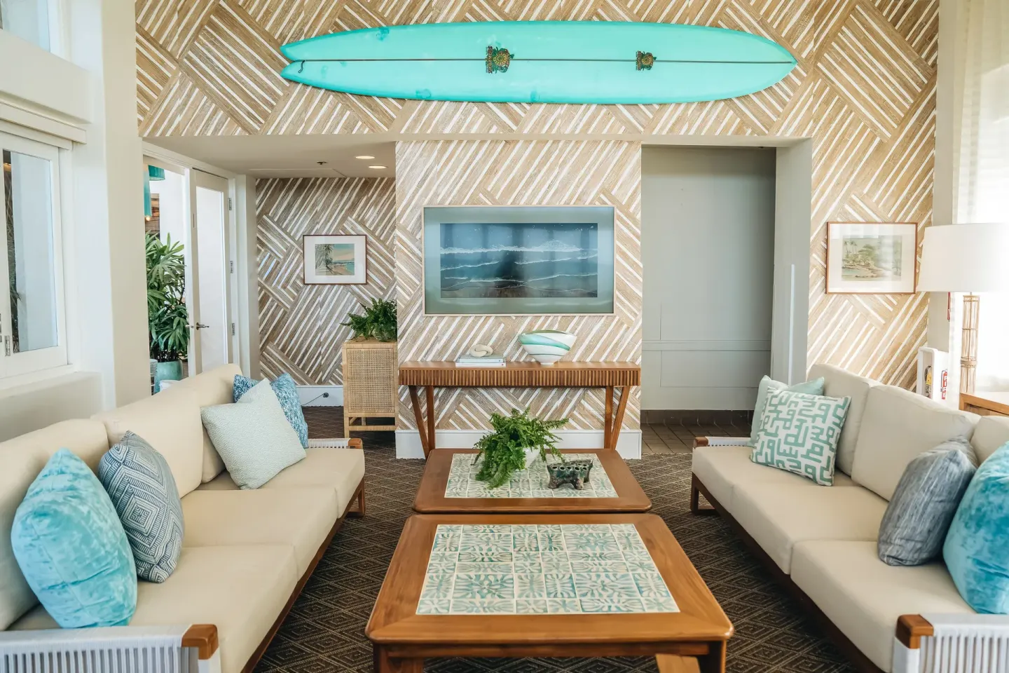 A lounge with sofas, patterned walls, and a turquoise surfboard is shown at Kaimana Beach Hotel.