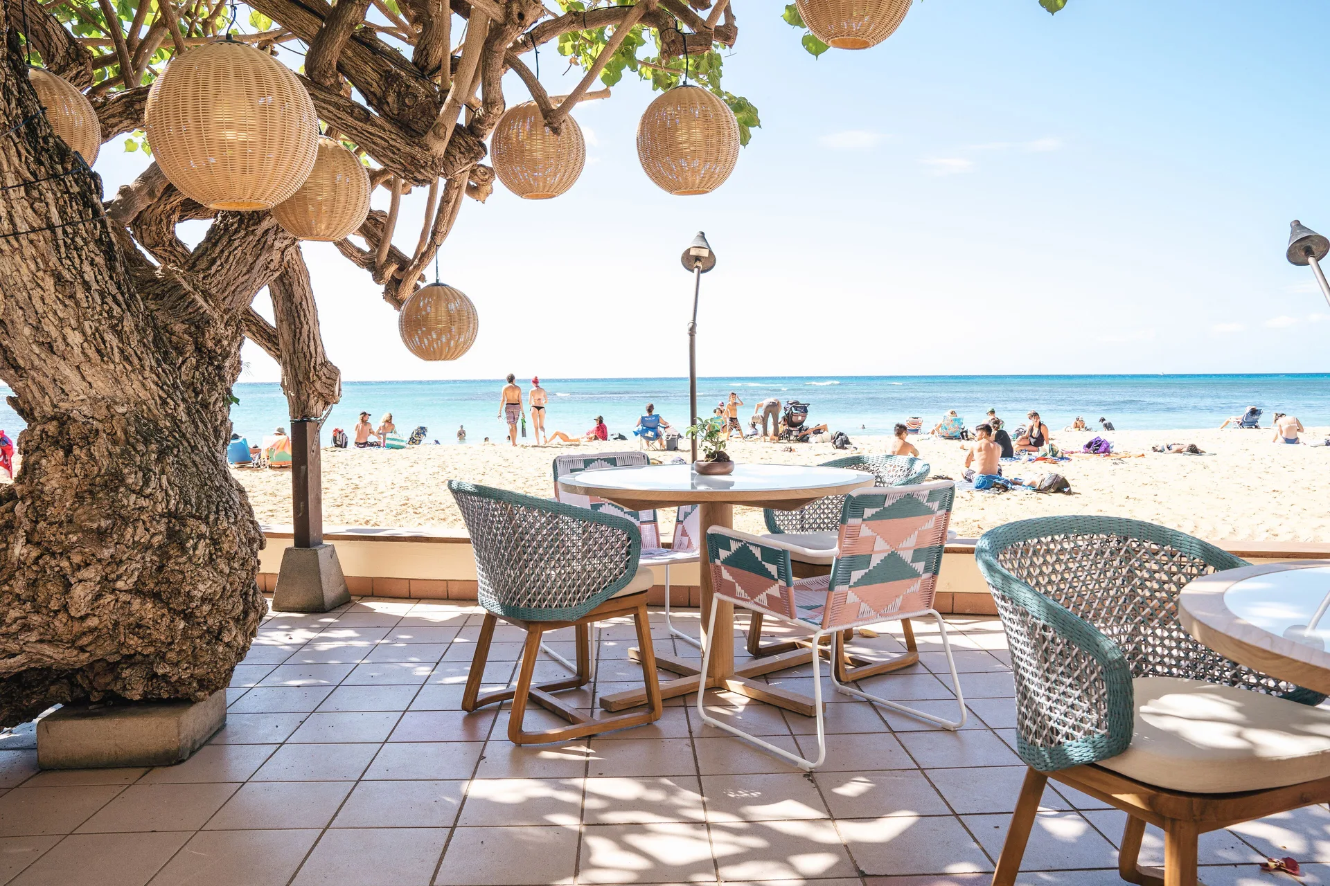 Beachfront patio tables sit under a tree with woven lanterns at Kaimana Beach Hotel.