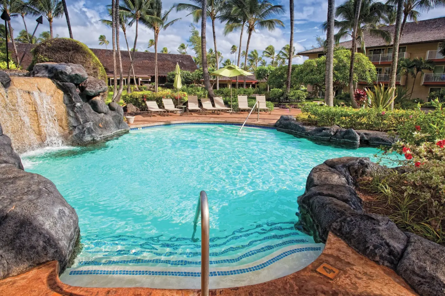 Swimming pool with rock waterfall, steps, lounge chairs, and palms at Kauai Coast at the Beachboy.