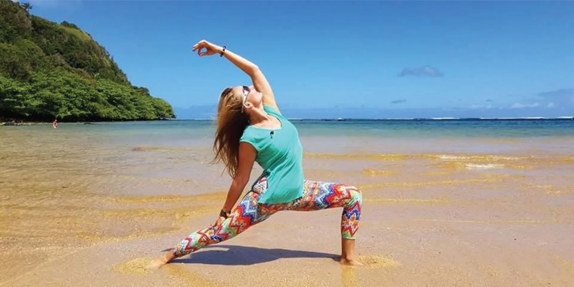 Person doing yoga on a sandy beach near Kauai Shores Hotel with ocean and green hillside.