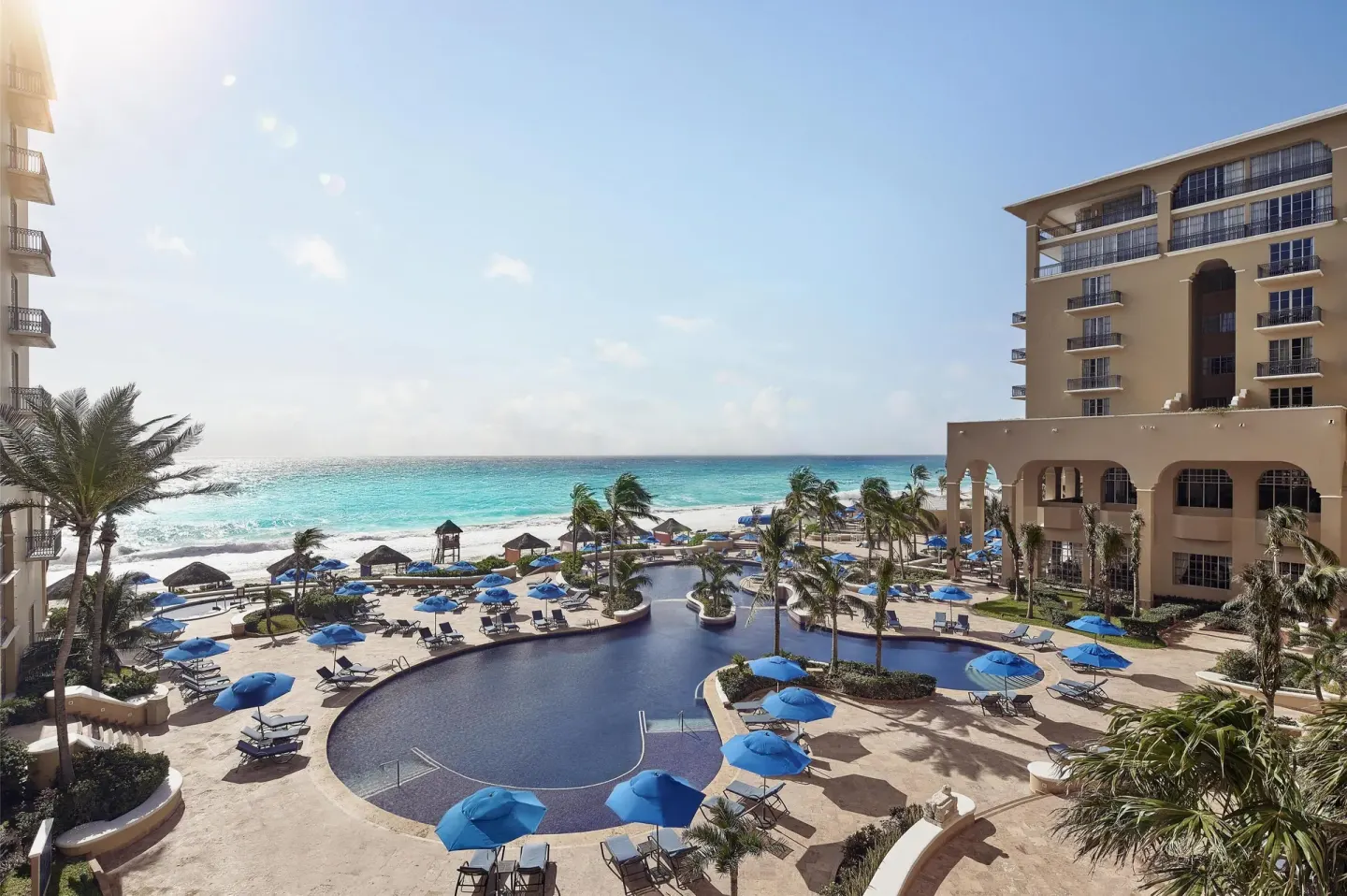 Resort pool courtyard with blue umbrellas and ocean beyond at Kempinski Hotel Cancún.