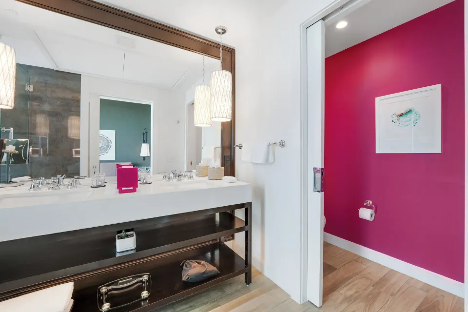 Bathroom vanity with mirror reflecting bedroom and pink-walled toilet room in Yacht Club View King Room at Kimpton Seafire Re
