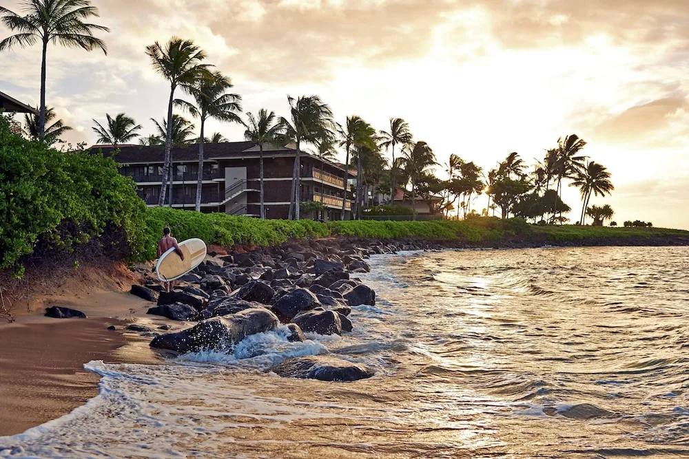 Surfer walking along the rocky shoreline at sunset by Ko'a Kea Resort on Po'ipu Beach.