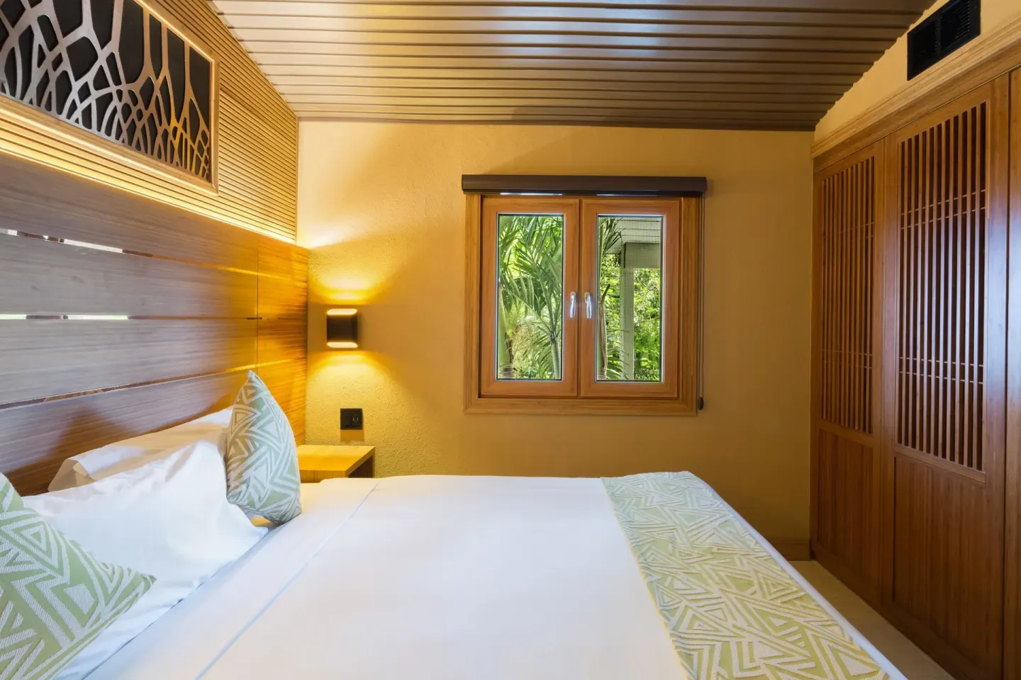 Guest room bed beside a window and wood wardrobe at Kokomo Botanical Resort.
