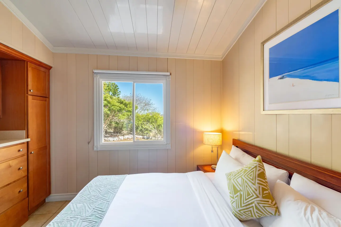 Guest room bed beside a wood cabinet and sunny window at Kokomo Botanical Resort.