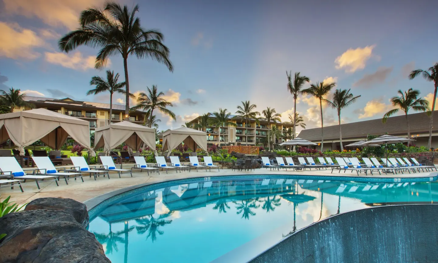 Pool deck with cabanas, lounge chairs, palms, and sunset sky at Koloa Landing Resort at Poipu.