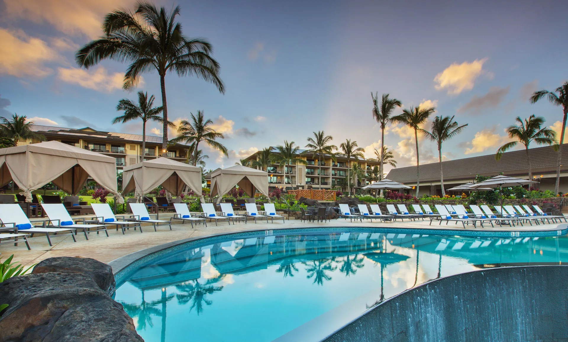 Pool deck with cabanas, lounge chairs, palms, and sunset sky at Koloa Landing Resort at Poipu.