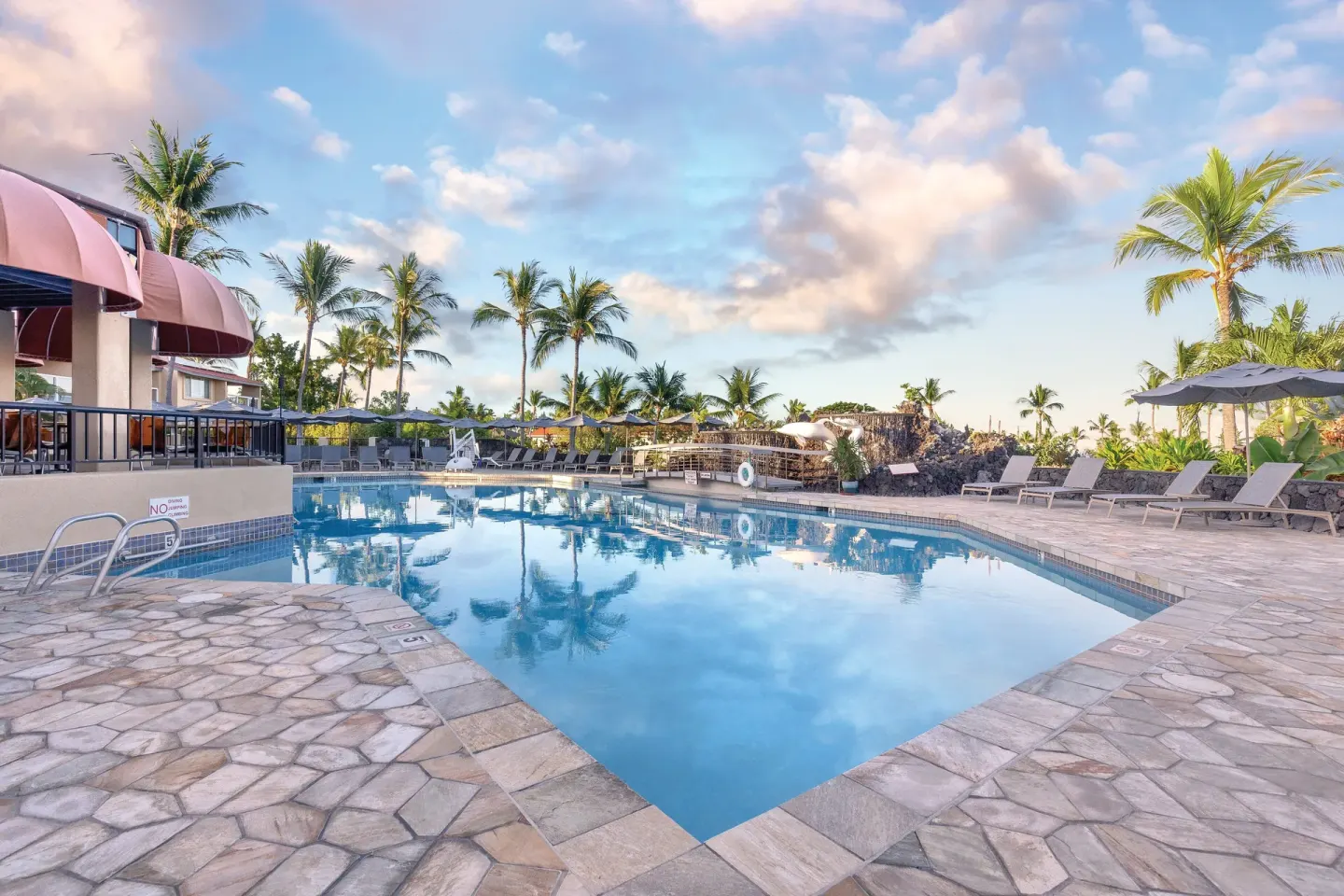 Large outdoor pool reflecting palms and sky beside loungers at Kona Coast Resort.