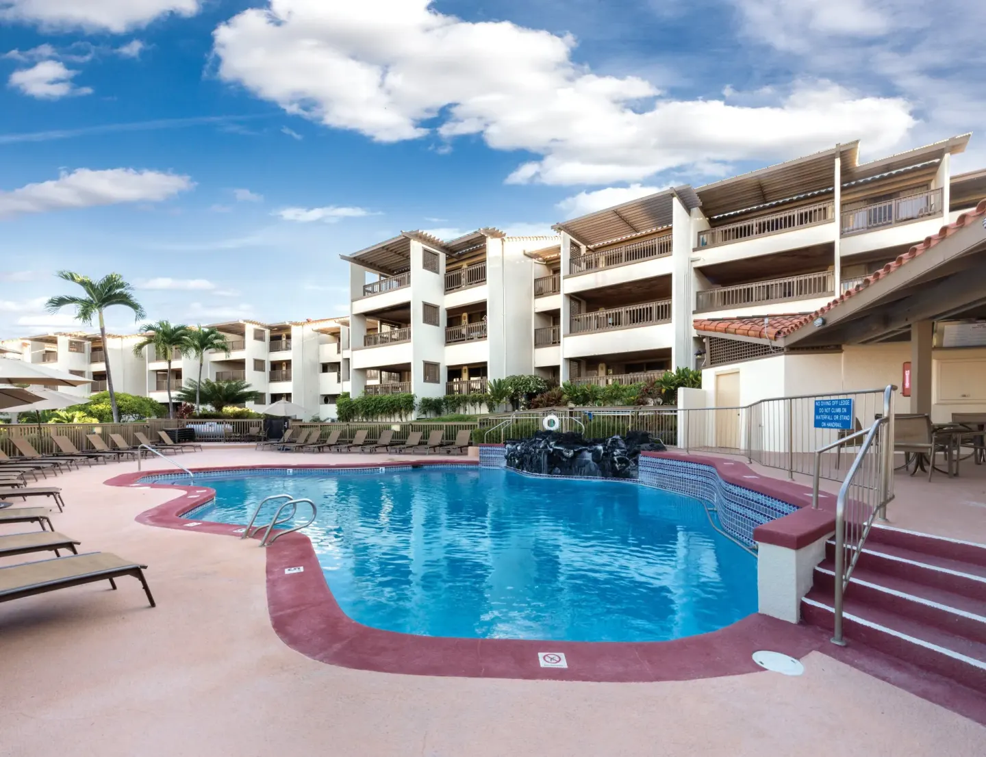 Pool courtyard with resort buildings, loungers, stairs, and water feature at Kona Coast Resort.