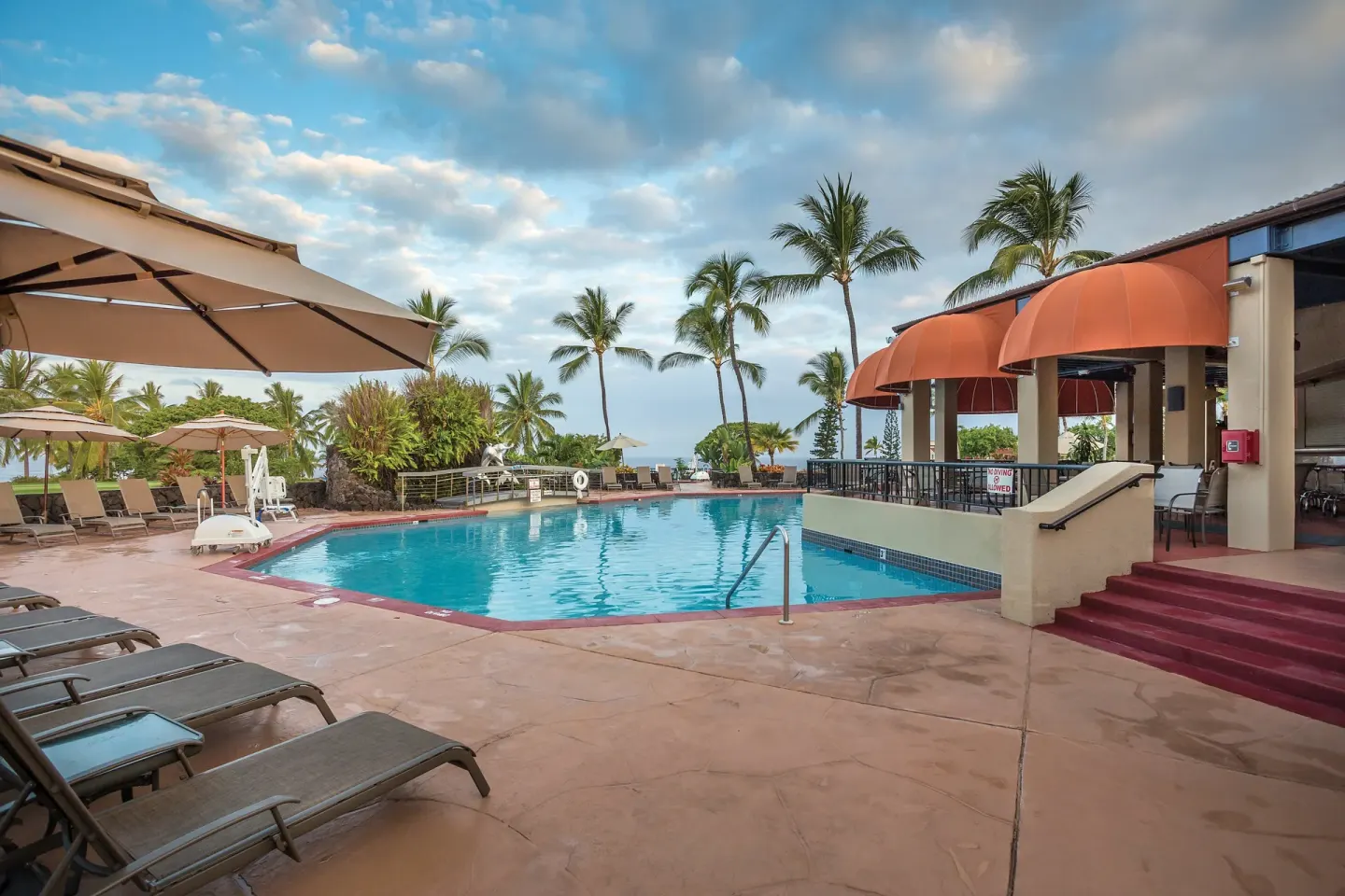 Swimming pool with lounge chairs, umbrellas, palms, and shaded building at Kona Coast Resort.