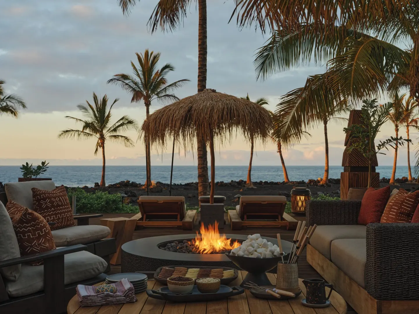 Beachfront fire pit lounge with s’mores setup at sunset at Kona Village.
