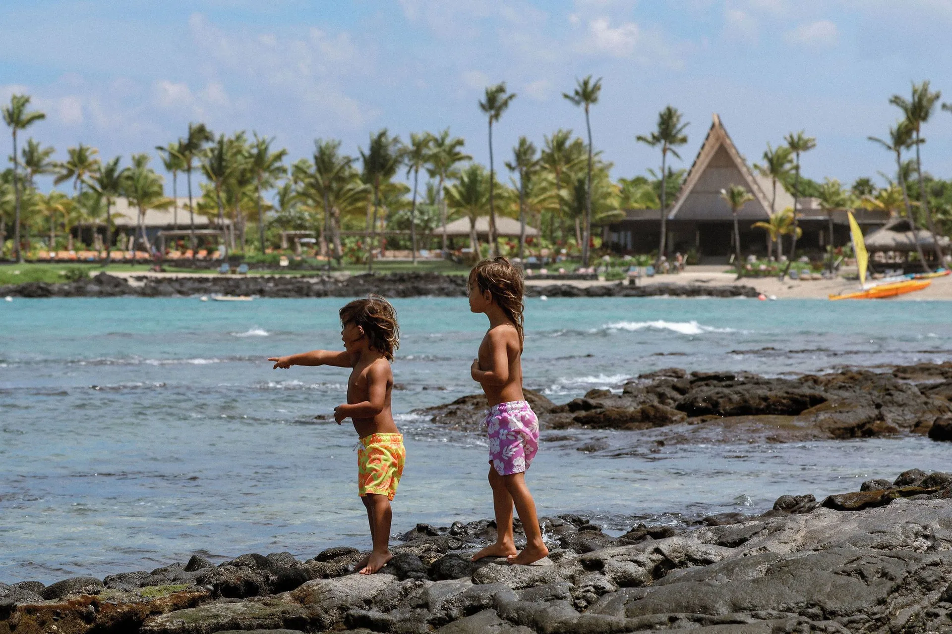 Two children stand on black lava rocks by the shoreline at Kona Village.