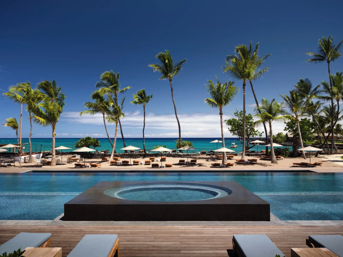 Pool deck with loungers, palms, beach umbrellas and ocean beyond at Kona Village.